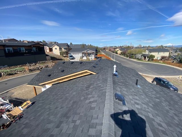 Rooftop view of a house with newly installed dark gray shingles, sunny day. Street and houses in the background.