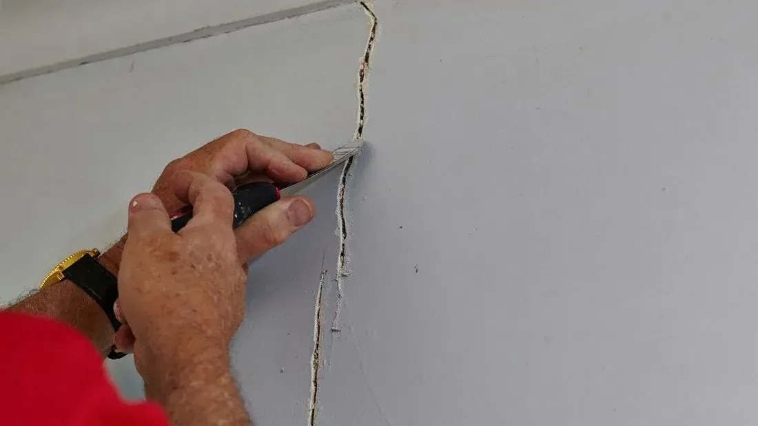 Person using a putty knife to scrape a crack in a light gray wall.