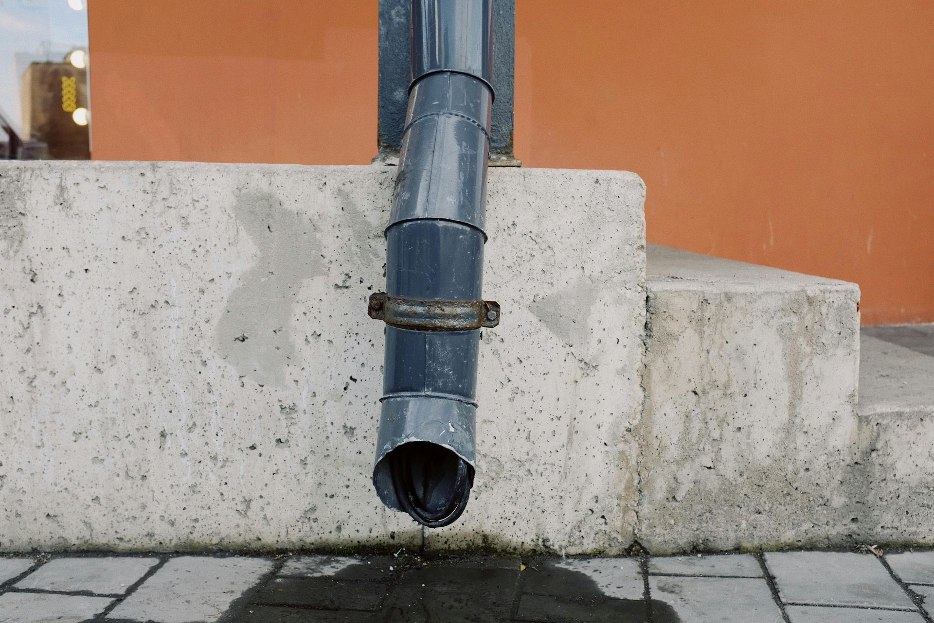 Rainwater flows from a dark gray downspout attached to a concrete structure; a brick wall is in the background.