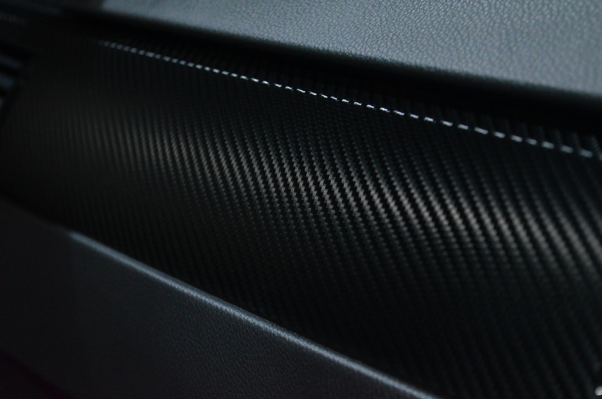 Close-up of a black carbon fiber textured car interior panel with white stitching.