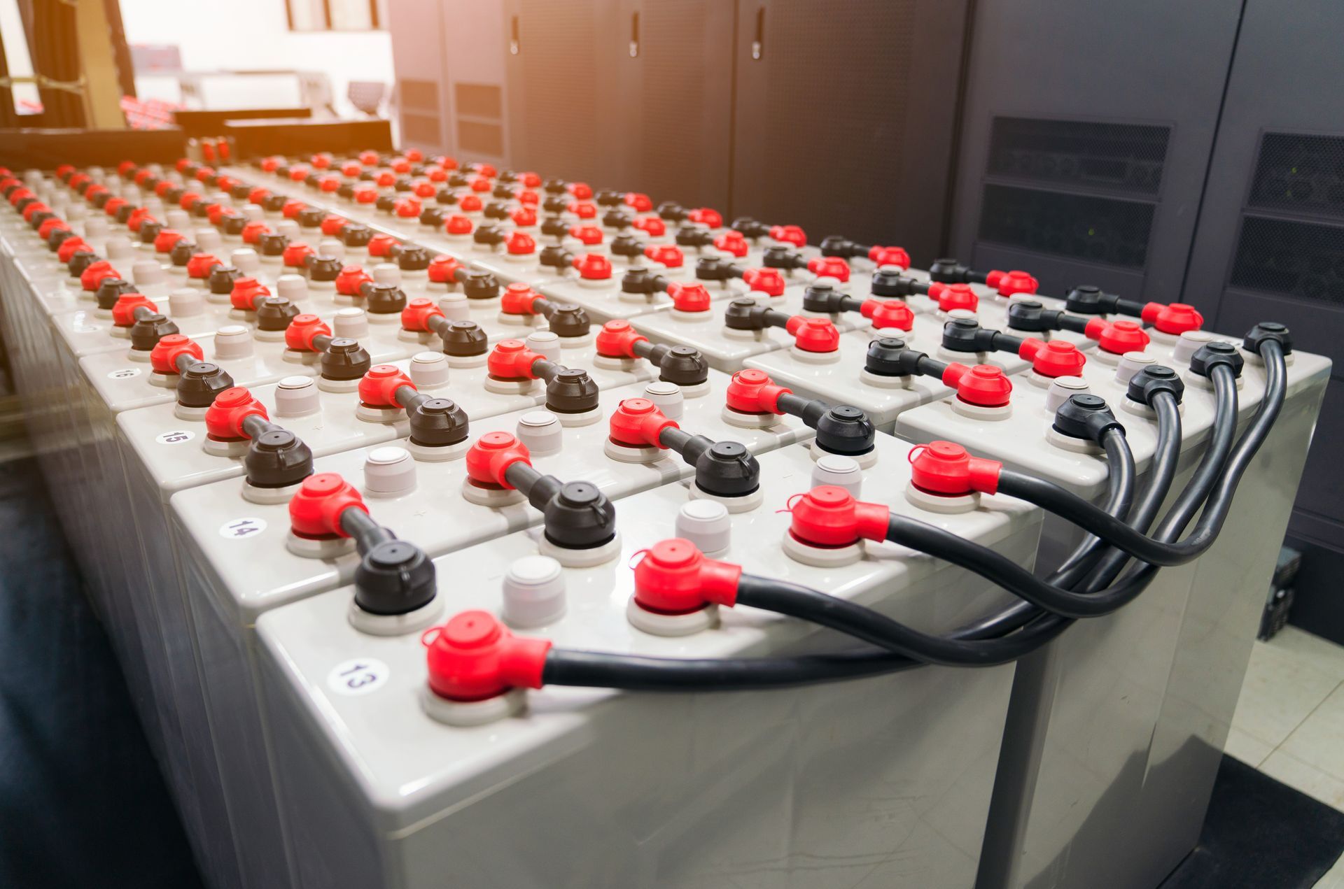 Rows of large batteries in a server room, red and black terminals connected by black cables.