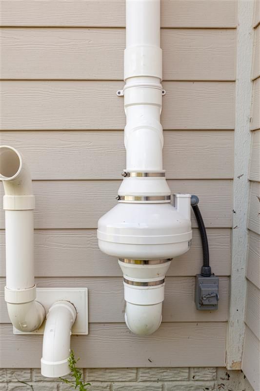 White PVC pipes and a round component attached to a beige siding wall; includes an electrical outlet.