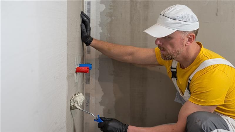 Person in yellow shirt painting a wall with a roller. Wearing a white cap and black gloves, indoor setting.