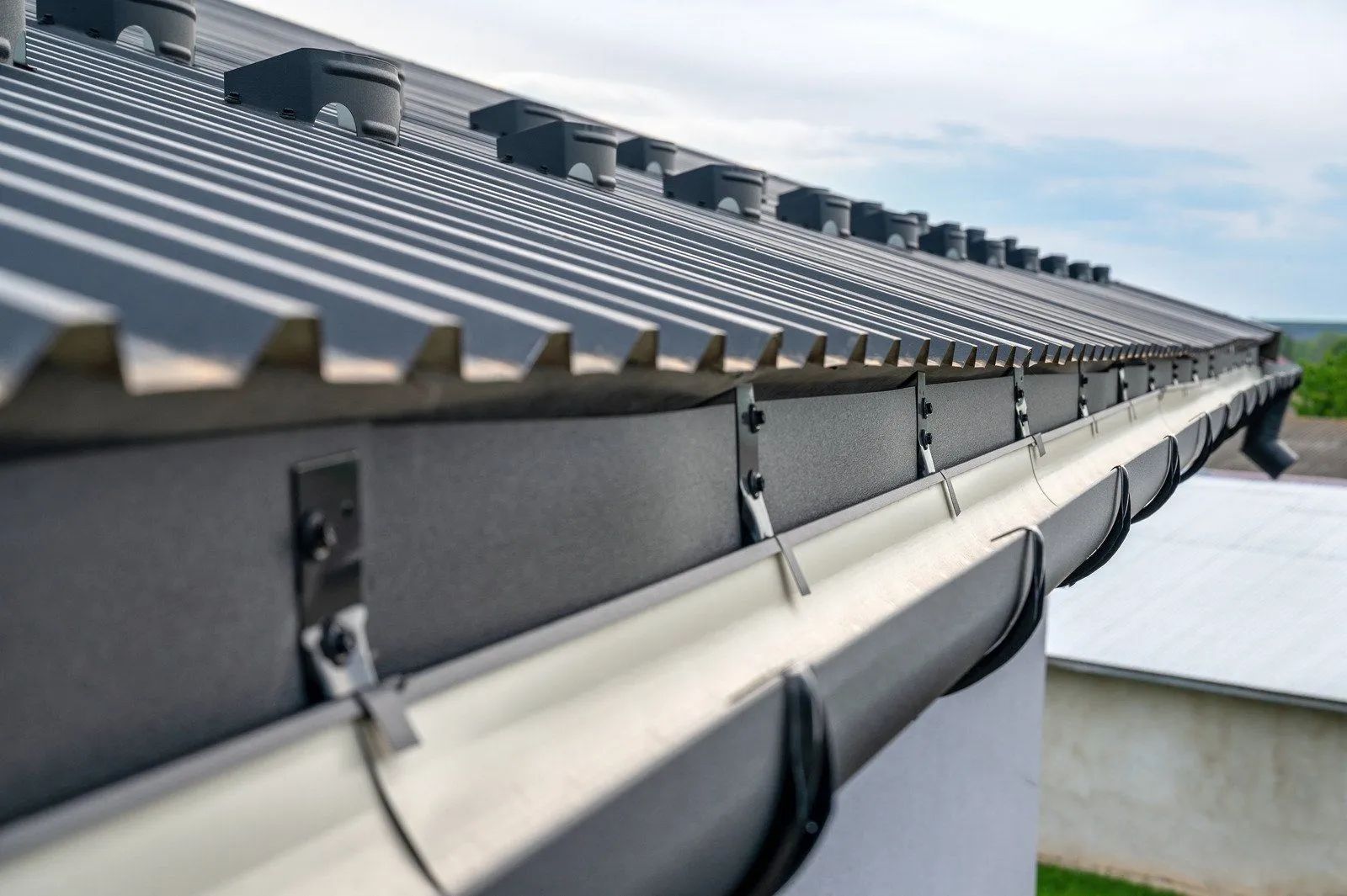 Row of gray metal roof panels and gutters on a sloped building under a cloudy sky