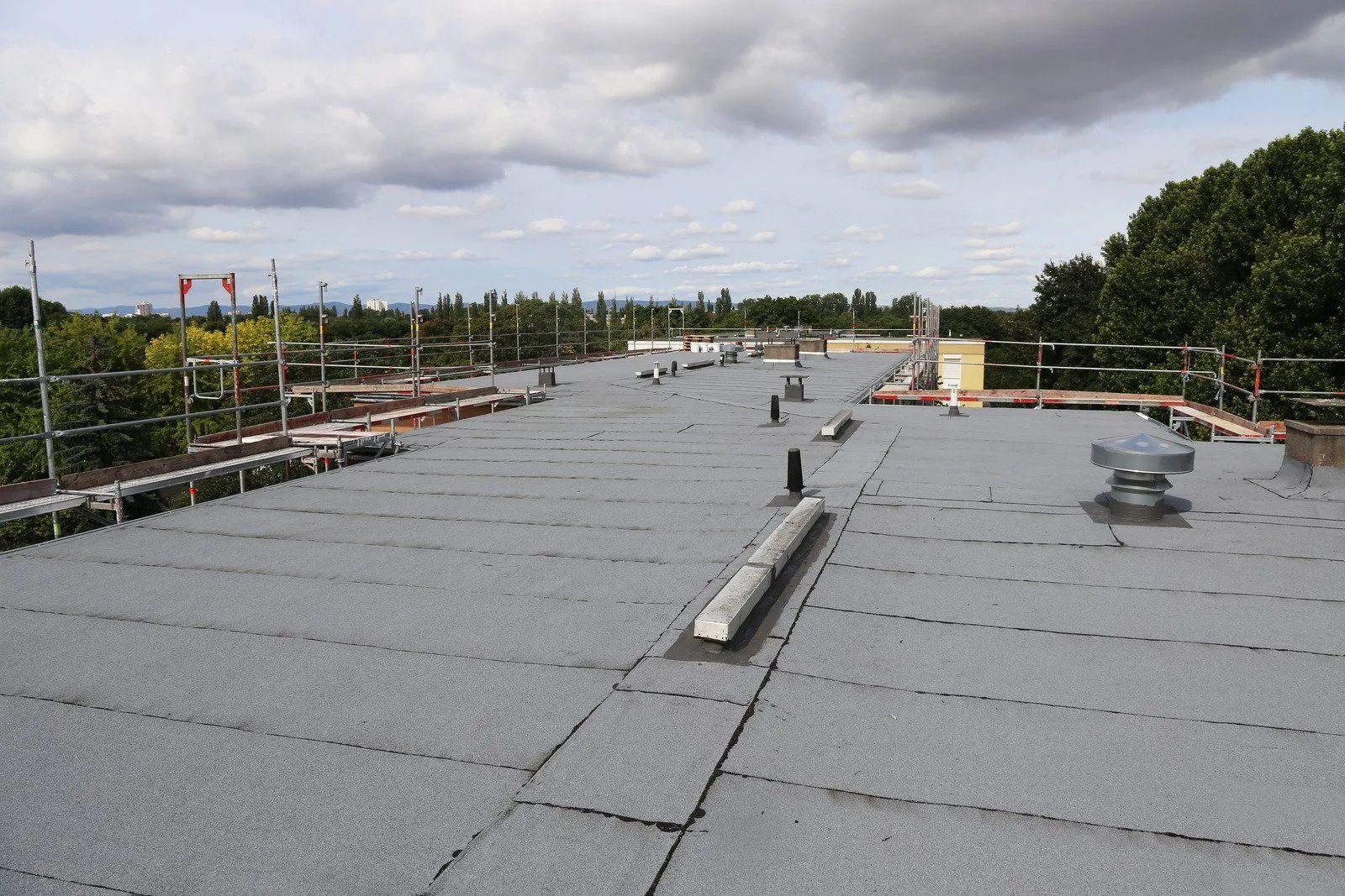 Flat, gray commercial roof with vents, surrounded by scaffolding and a cloudy sky.