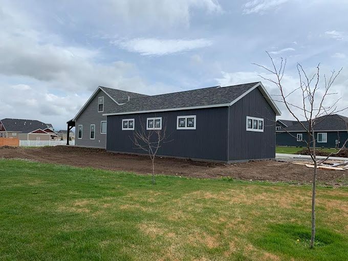 Back of a dark gray house with a light gray side, windows, and a grassy lawn under a cloudy sky.