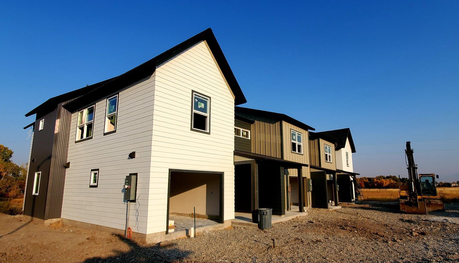 New townhomes under construction, white and dark siding, blue sky, gravel lot.