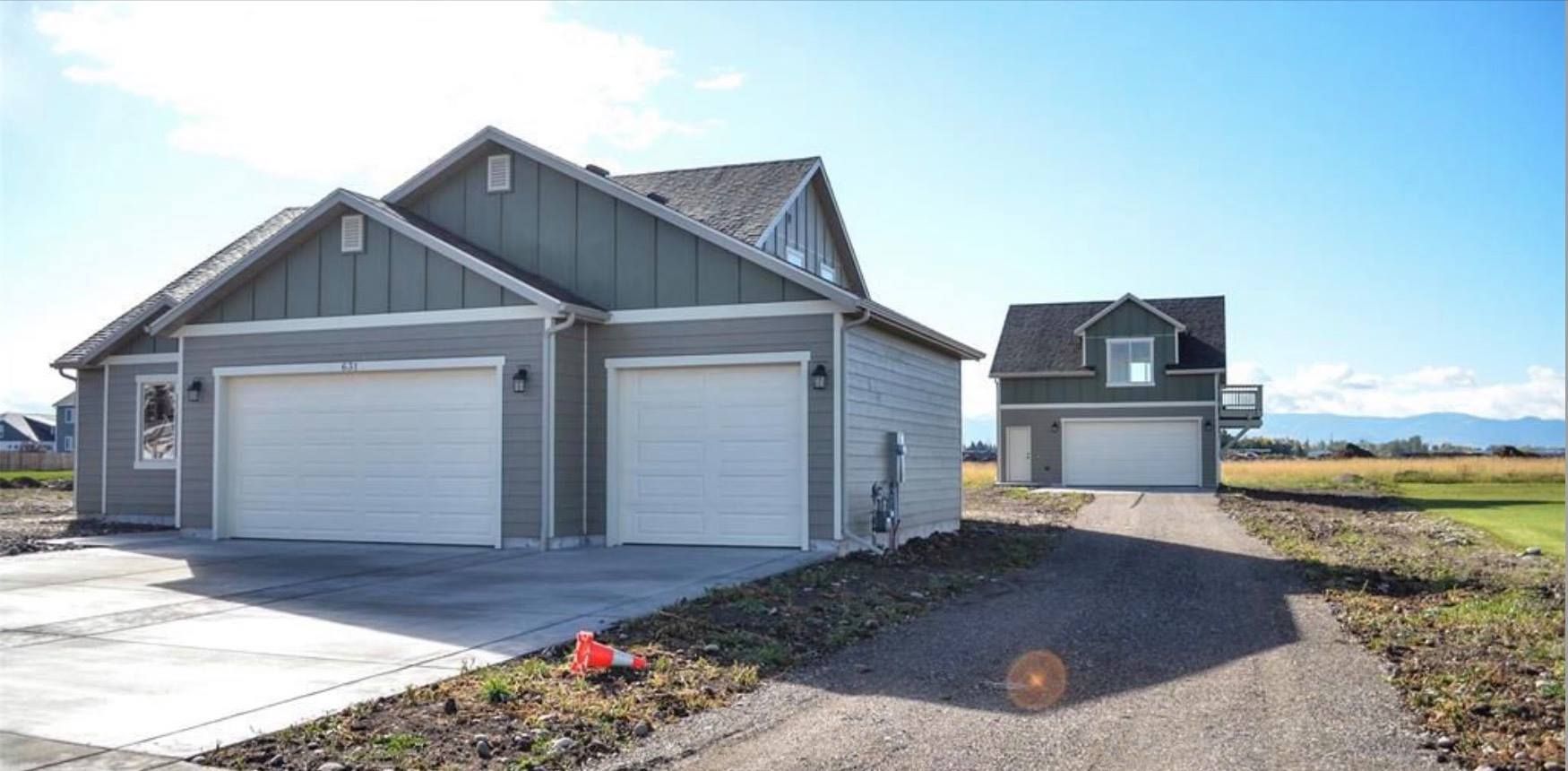Two-car garage with attached house and detached building with garage, on a gravel drive, field in background.