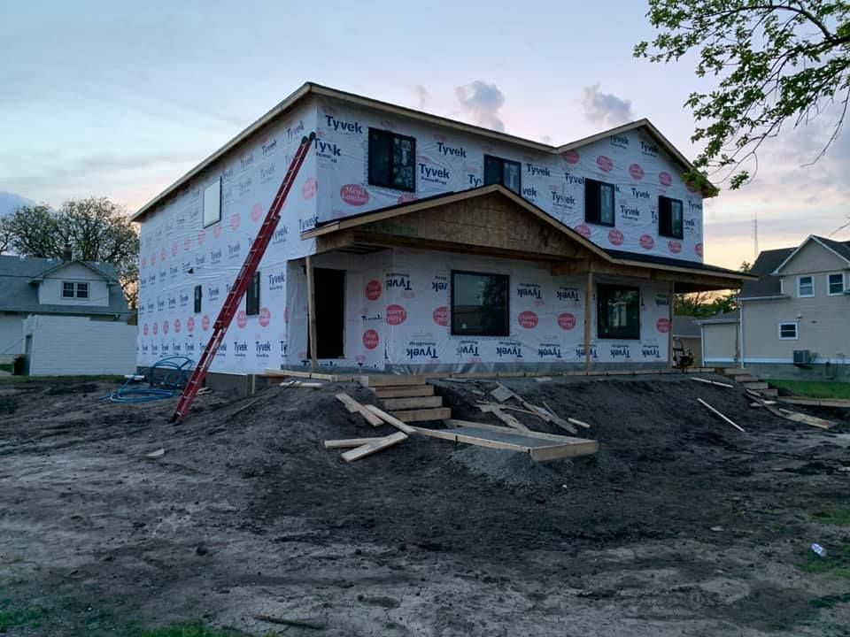 House under construction, covered in Tyvek, with a ladder leaning against it. Dirt mound in front.