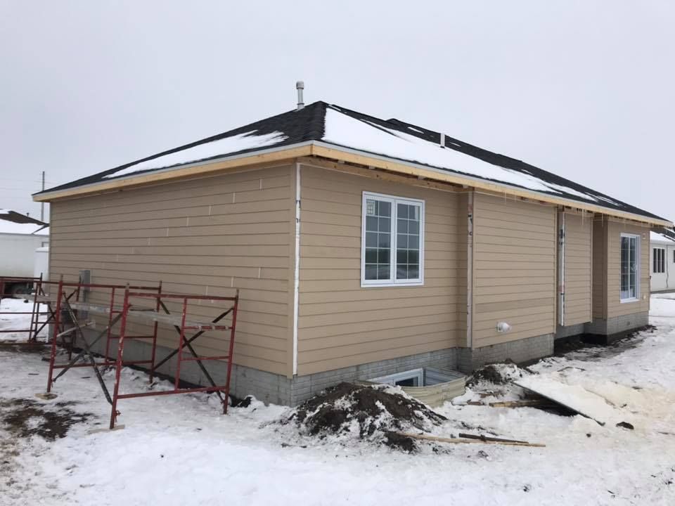 Tan-sided house under construction with white trim, windows, and black roof. Snow covers the ground.