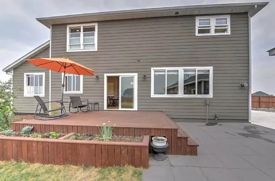 Back of a two-story house with a wooden deck, orange umbrella, and landscaping against a cloudy sky.