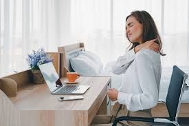Woman sitting at a desk, holding her shoulder while working on a laptop in a bright room.