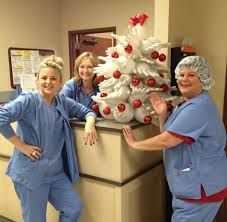 Three medical workers pose by a Christmas tree made of white gloves, decorated with ornaments.