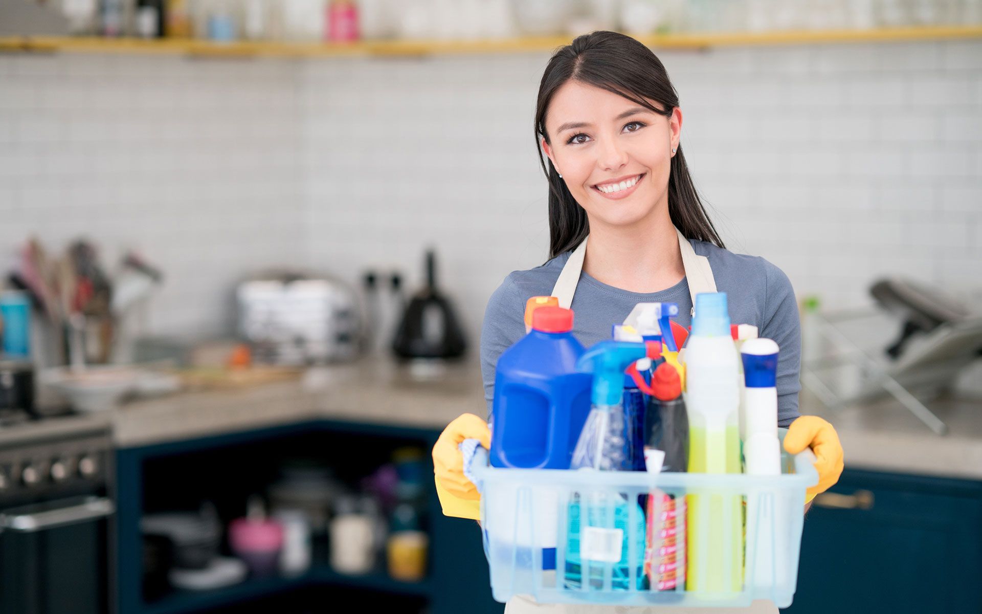 A woman is holding a basket full of cleaning supplies in a kitchen.