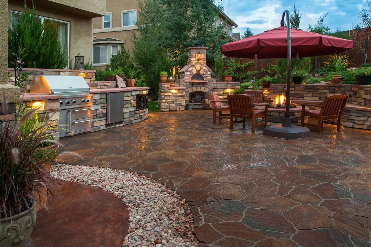 Backyard patio with outdoor kitchen, fireplace, seating, and umbrella; brown and red tones.