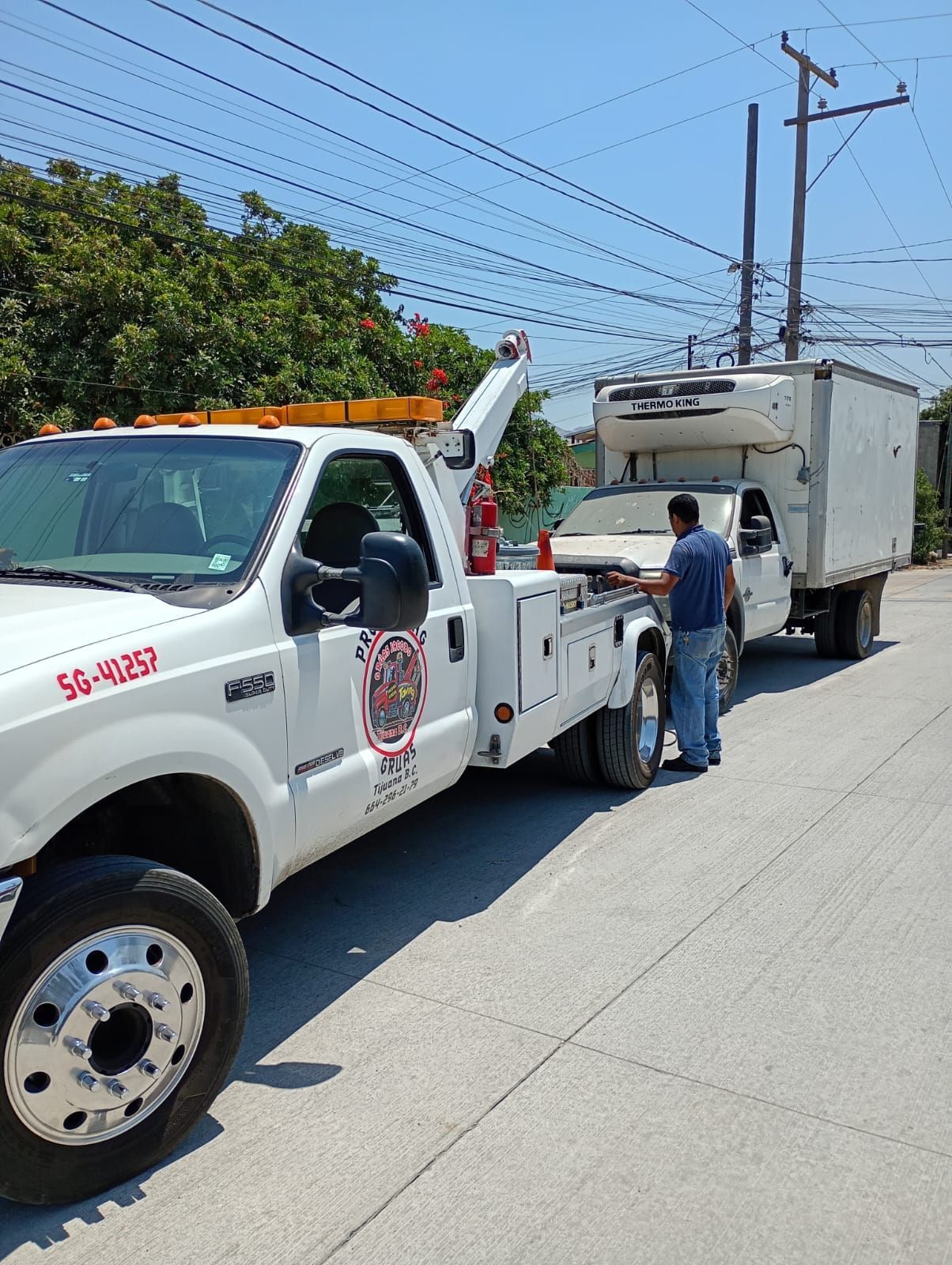Una grúa blanca está enganchada a un camión frigorífico en una carretera asfaltada bajo un cielo azul brillante.