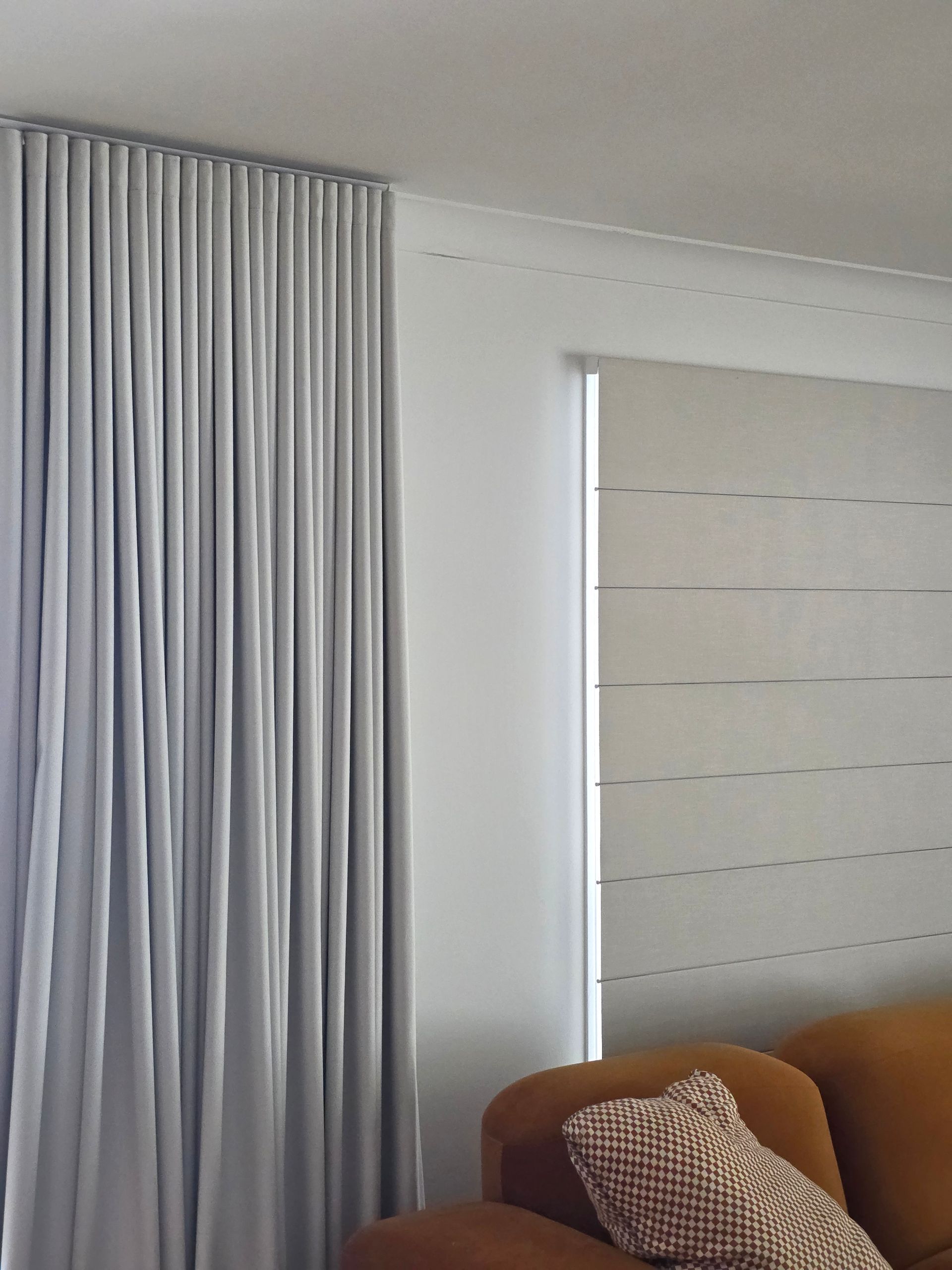 White curtains and a window blind beside a brown sofa with a patterned pillow in a bright room