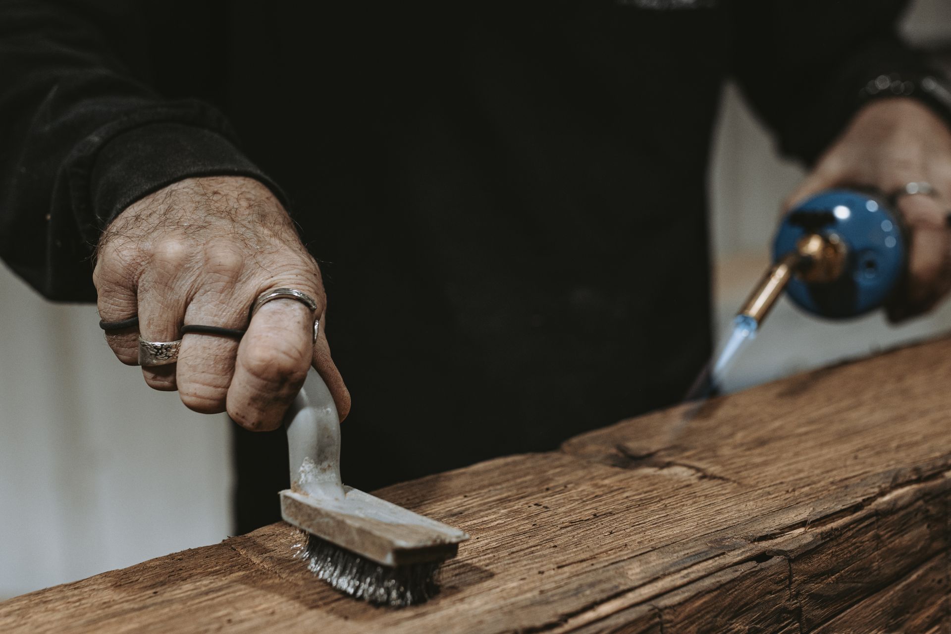 Hands using a blowtorch and wire brush for historic barn restoration on aged wood.