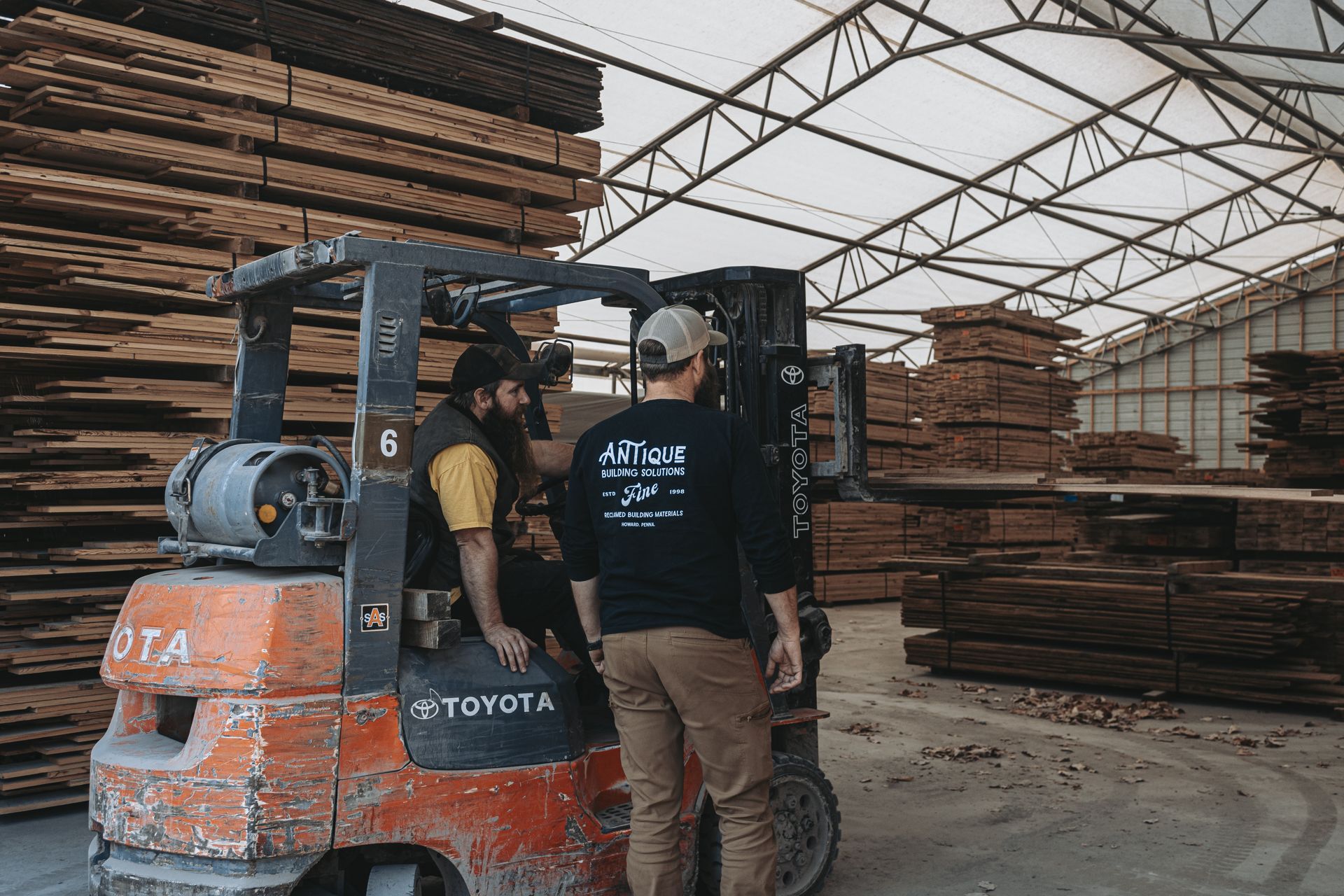 Two workers of restoration companies in a lumber warehouse filled with stacked wooden planks.