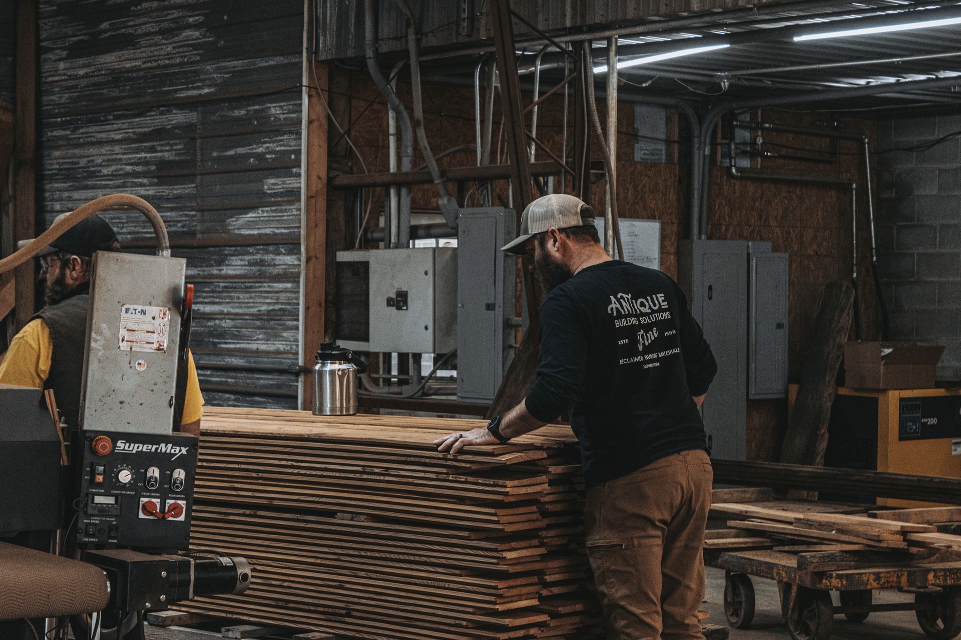 Two people working in a woodworking workshop with machinery, high-quality barn frames, and industrial equipment.