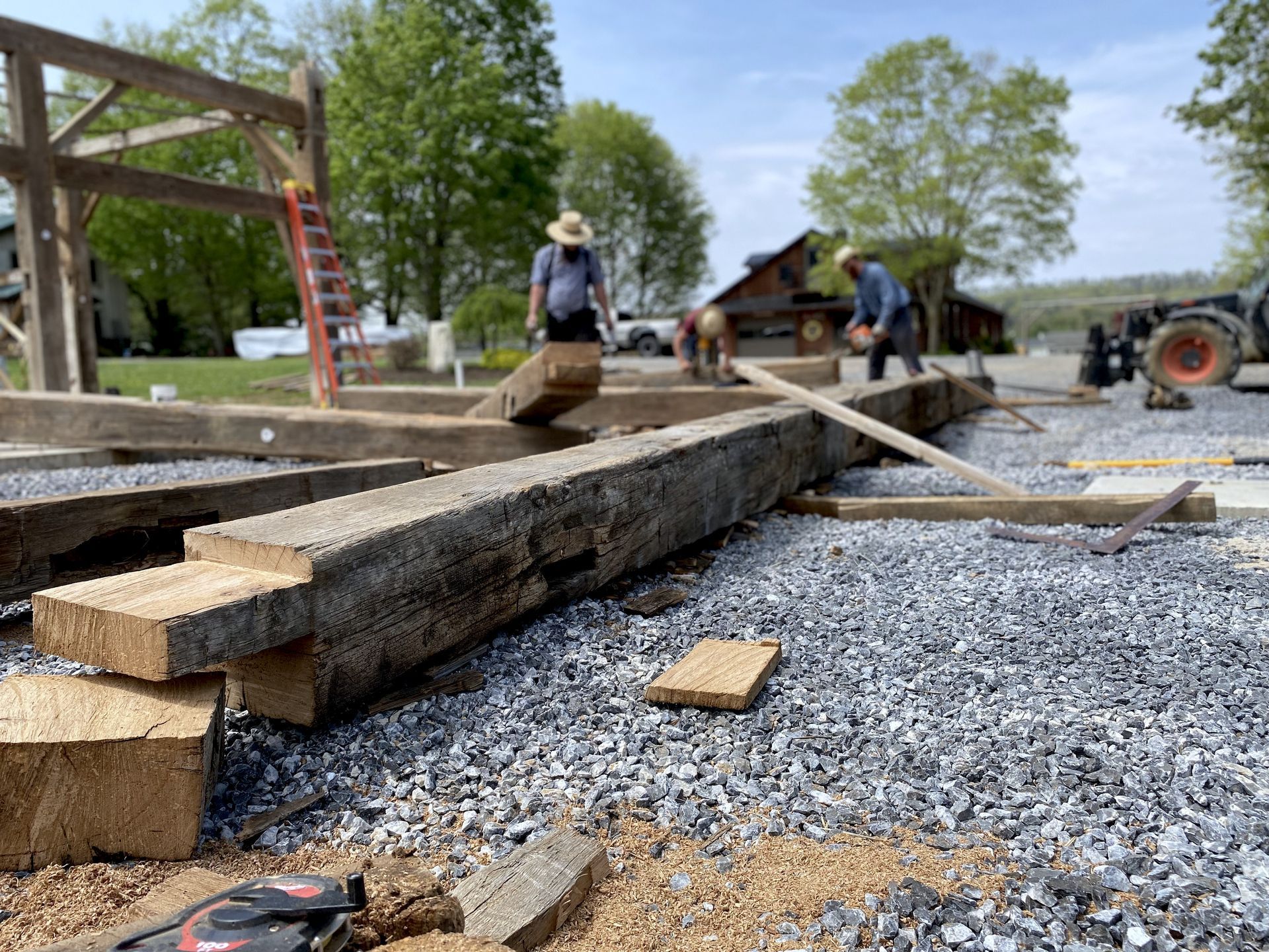 Workers shaping timber beams for barn framing on a rural site.