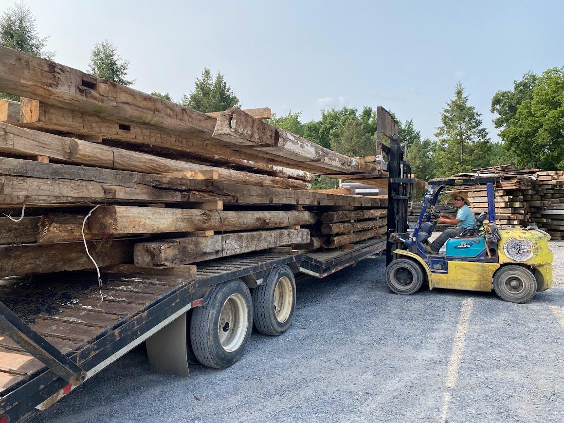 Forklift loading reclaimed wooden beams onto a trailer in a lumber yard, showcasing sourcing historic barn structures.