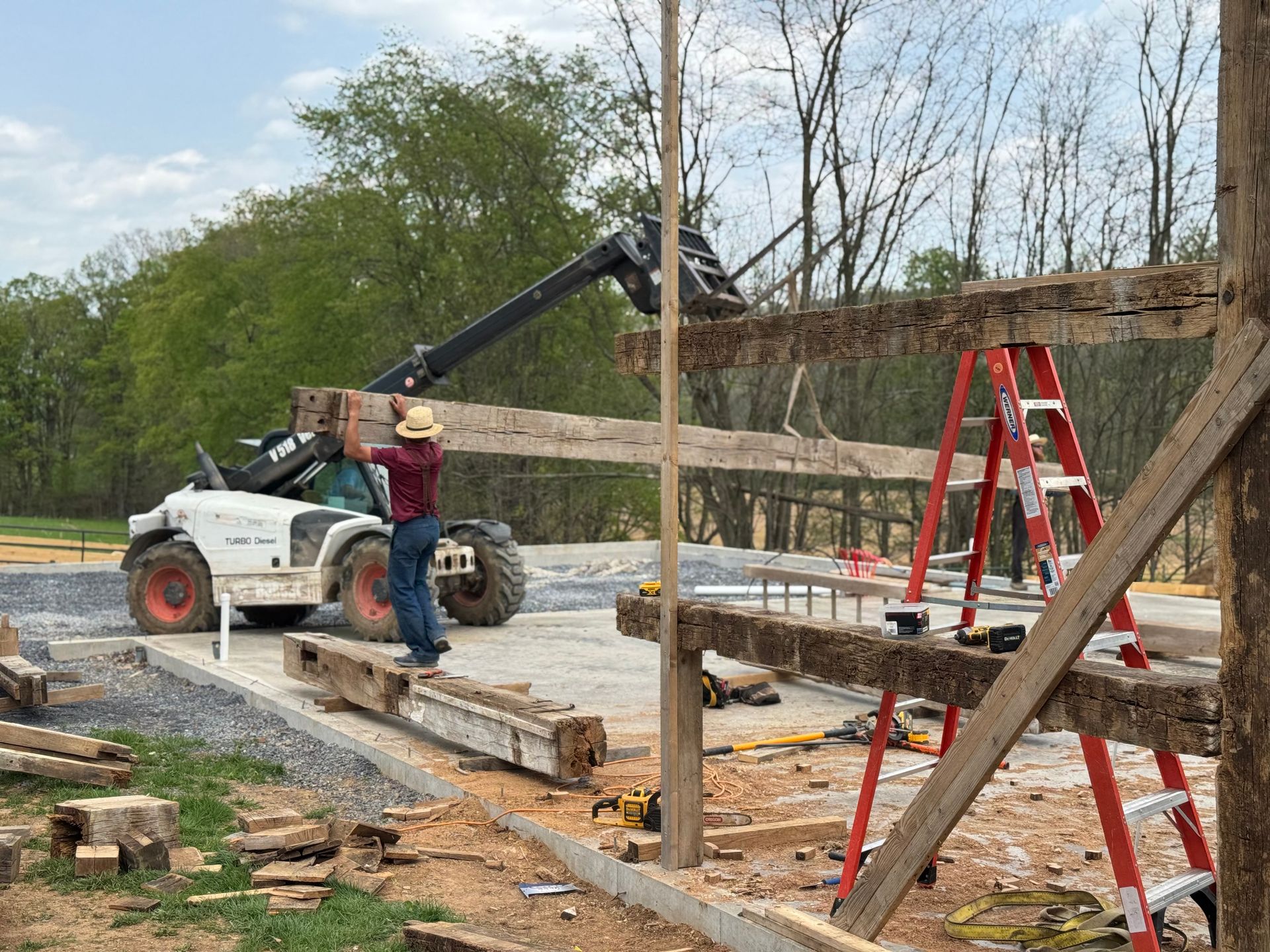 A worker guides a wooden beam being lifted by a telehandler as part of timber frame barn raising events.