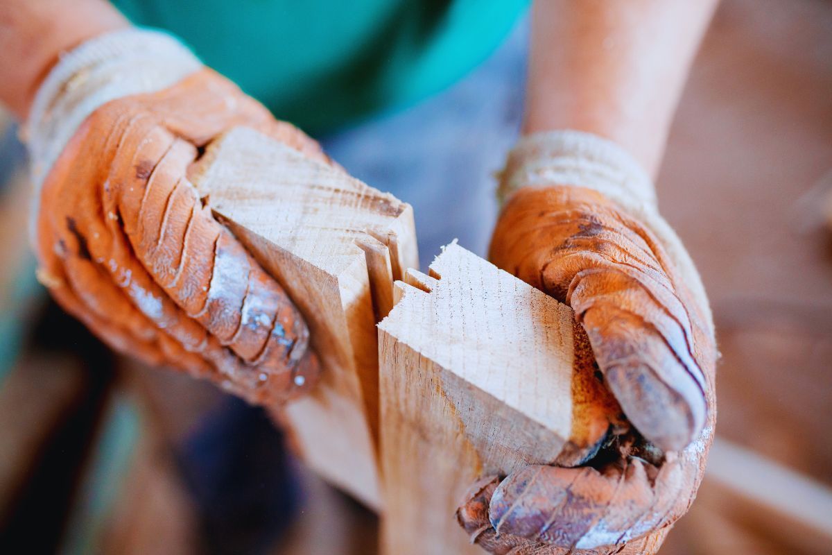 A close-up of gloved hands aligning two pieces of light-colored wood with interlocking cuts, showcasing traditional timber joinery techniques in a workshop setting.