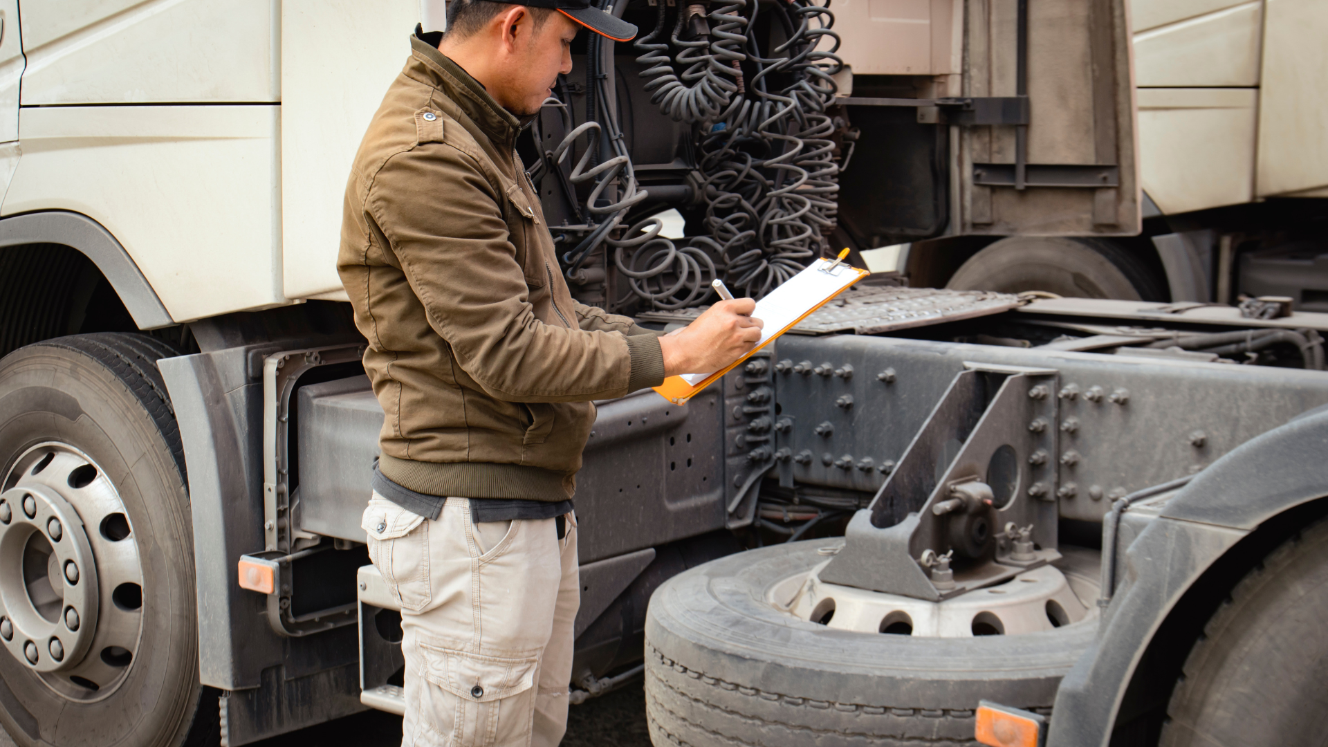 A person wearing a brown jacket and baseball cap stands by a semi-truck, filling out a document on a clipboard.