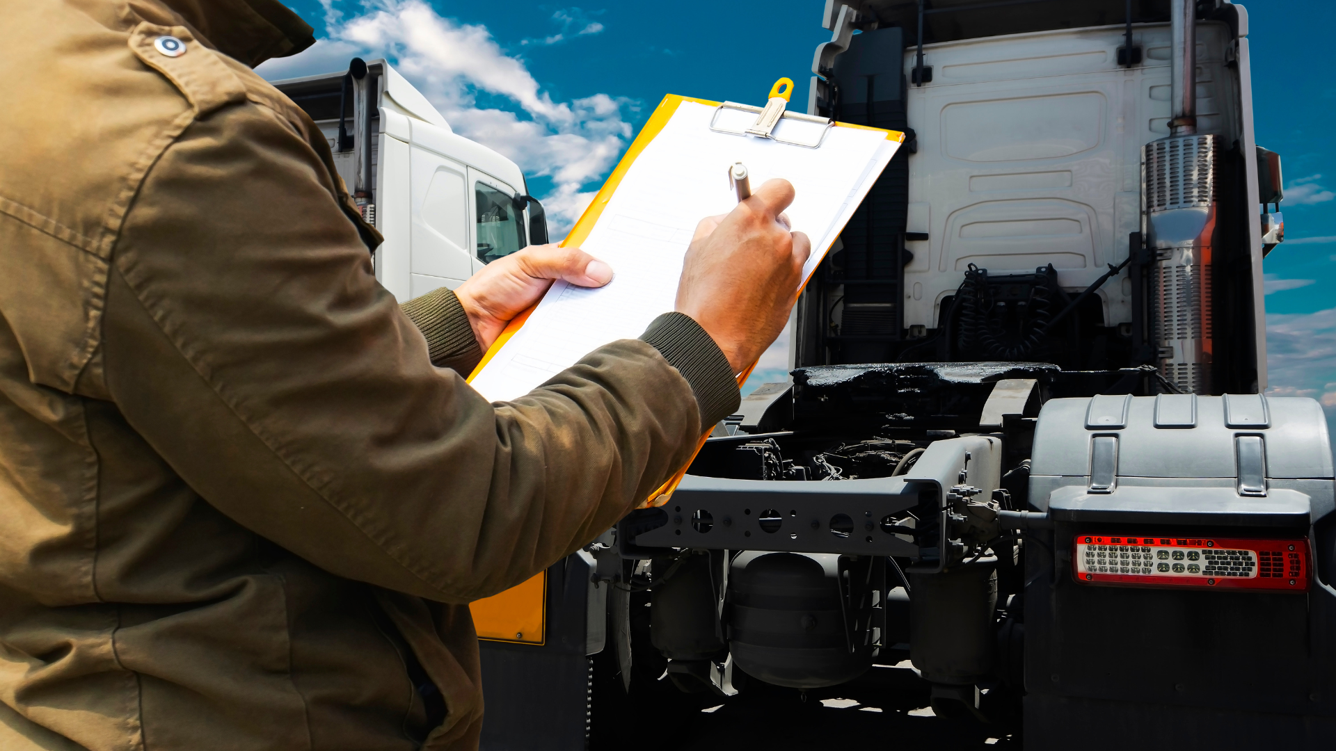 A person in a brown jacket writing on a clipboard in front of a semi-truck under a blue sky.