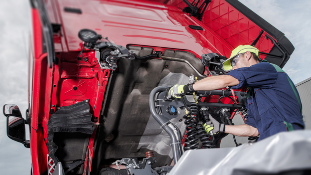 A mechanic in a yellow cap and work gloves repairs the exposed engine area of a large red truck cab.