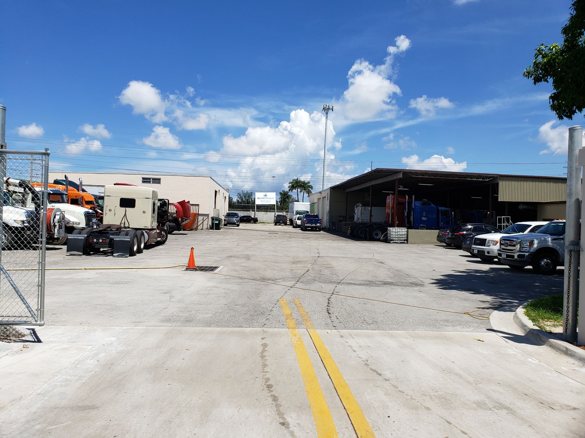A gated gravel truck lot with parked semi-trucks, a warehouse building, and vehicles under a sunny, partly cloudy sky.