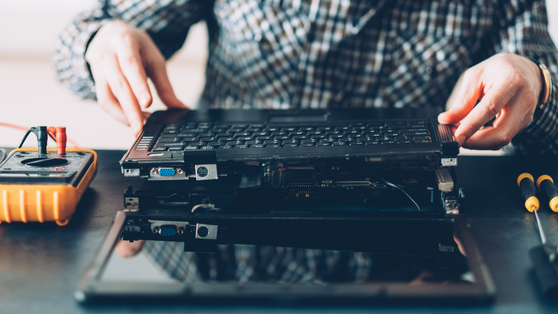 A person in a plaid shirt disassembles a laptop on a desk with a multimeter and screwdrivers nearby.