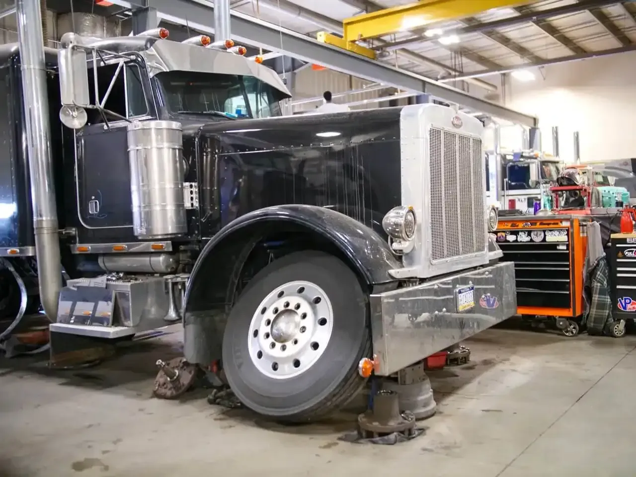 A black semi-truck parked in a garage with its front wheel removed and supported by jack stands.