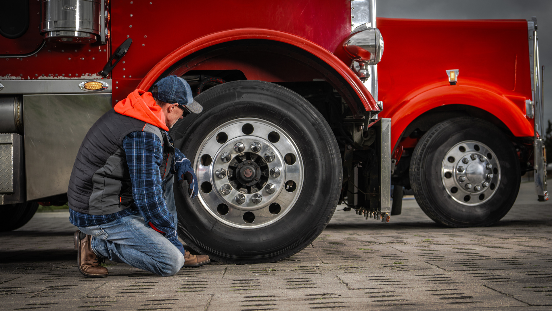 A person in work clothes kneels to inspect the wheel of a large, bright red semi-truck parked on a paved lot.