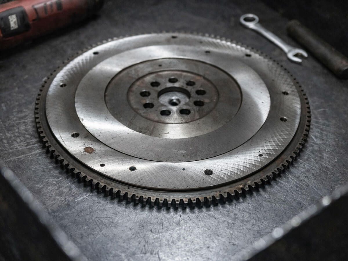 A metal engine flywheel resting on a dark, textured workbench next to a wrench and a tool handle.
