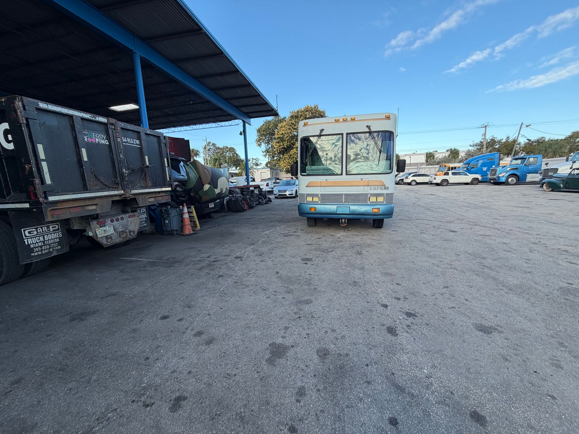 A white step van parked on a gravel lot under a covered bay next to a dark dump truck, with other vehicles in the distance.