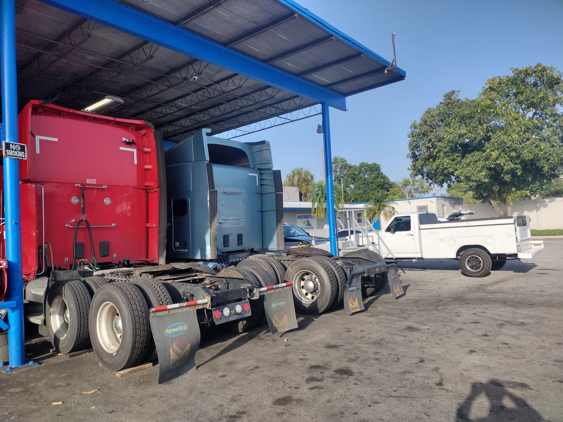 A red and a blue semi-truck cab parked under a blue metal canopy next to a white pickup truck on a paved lot.