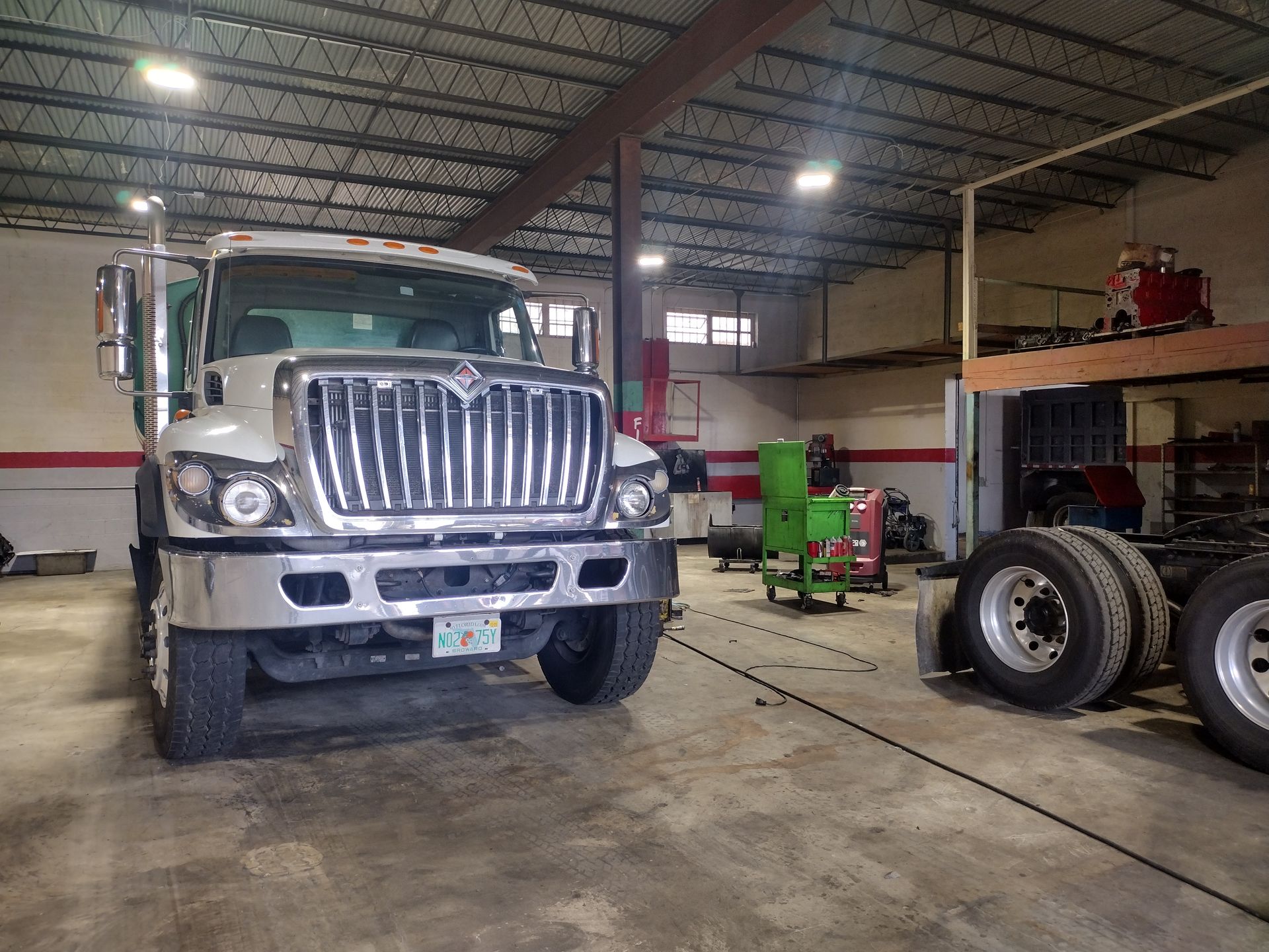 A white semi-truck parked inside a large, industrial garage with concrete floors and a lofted storage area.