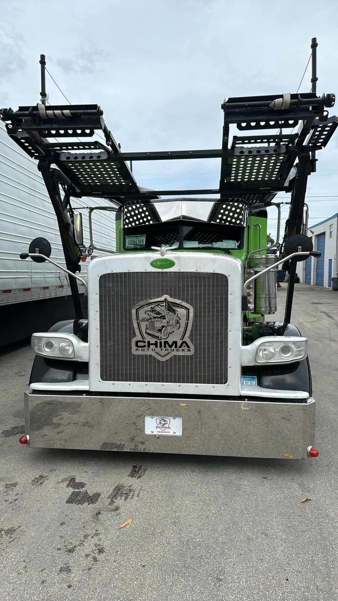 A green and white semi-truck auto hauler parked in an outdoor lot, featuring a large, customized shield-shaped hood emblem.