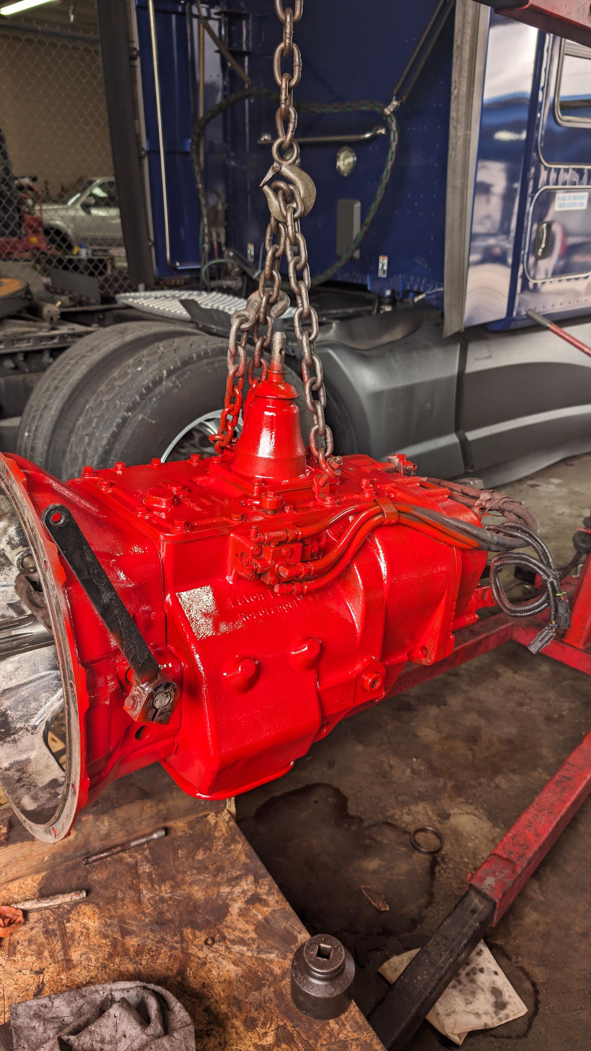 A freshly painted, bright red semi-truck transmission hangs from a chain hoist in a garage near a parked tractor-trailer.
