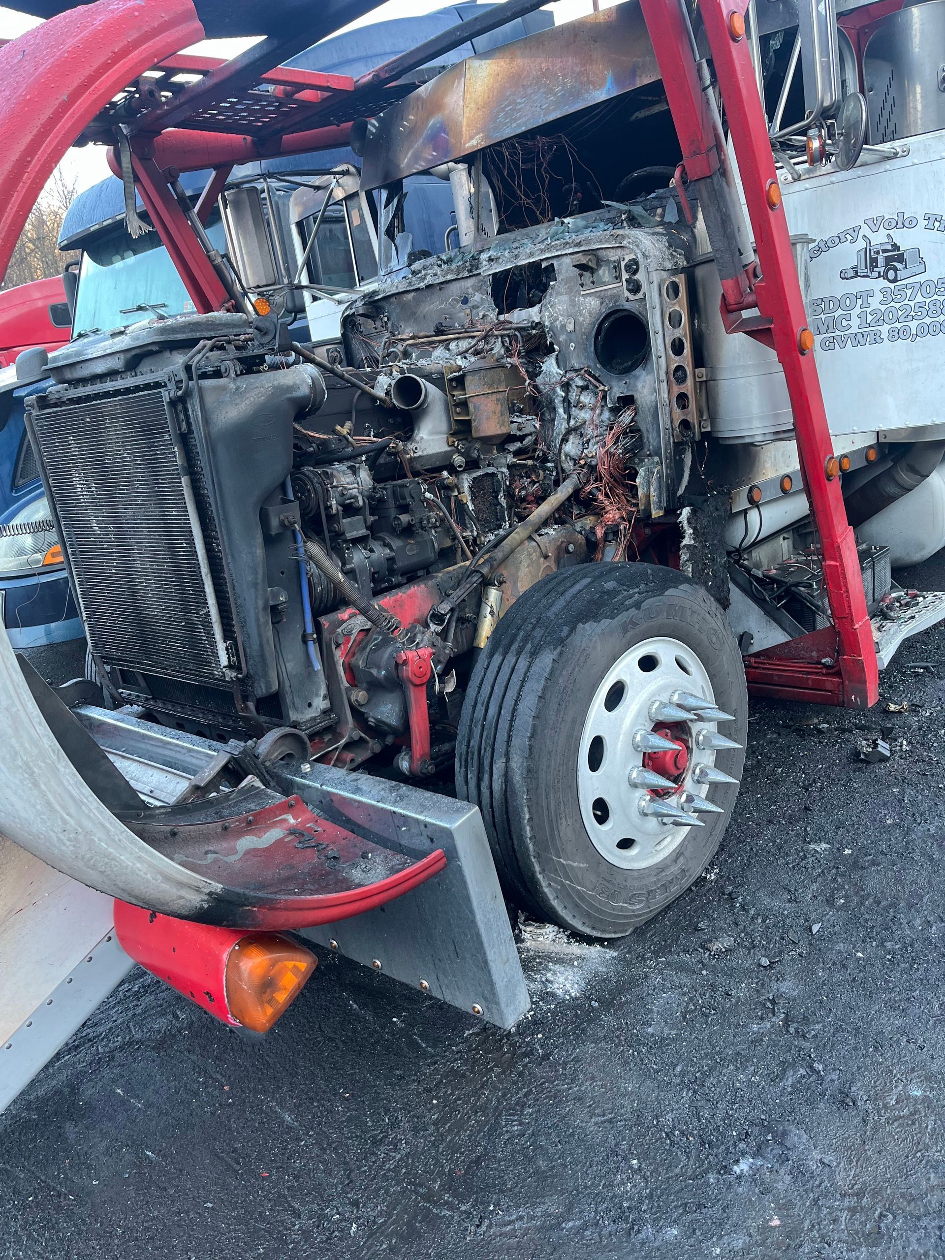 A heavily fire-damaged truck chassis with a charred engine, exposed radiator, and red frame parked on gravel.
