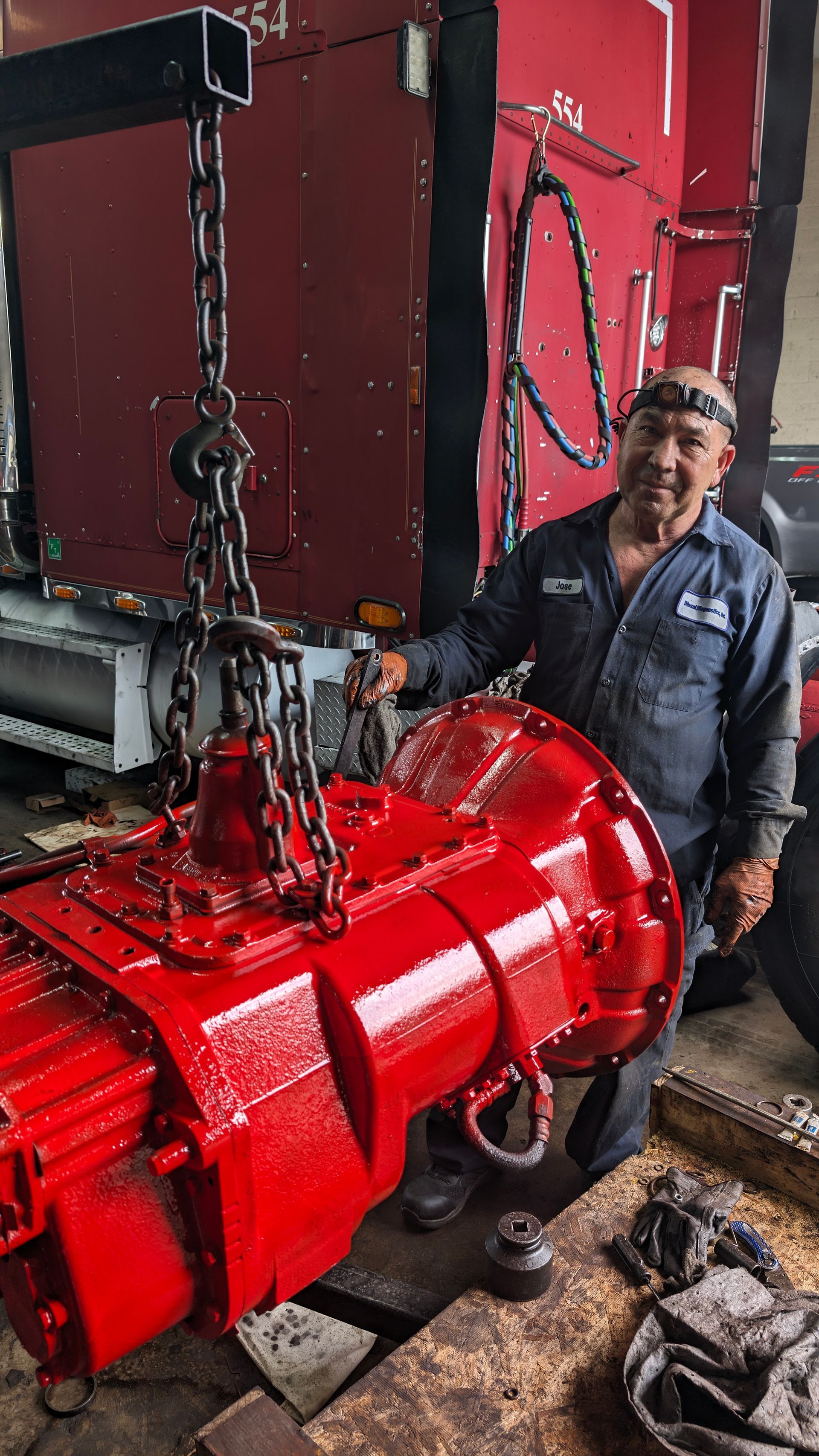 A mechanic stands next to a freshly painted, bright red automotive transmission suspended by a hoist in a garage.