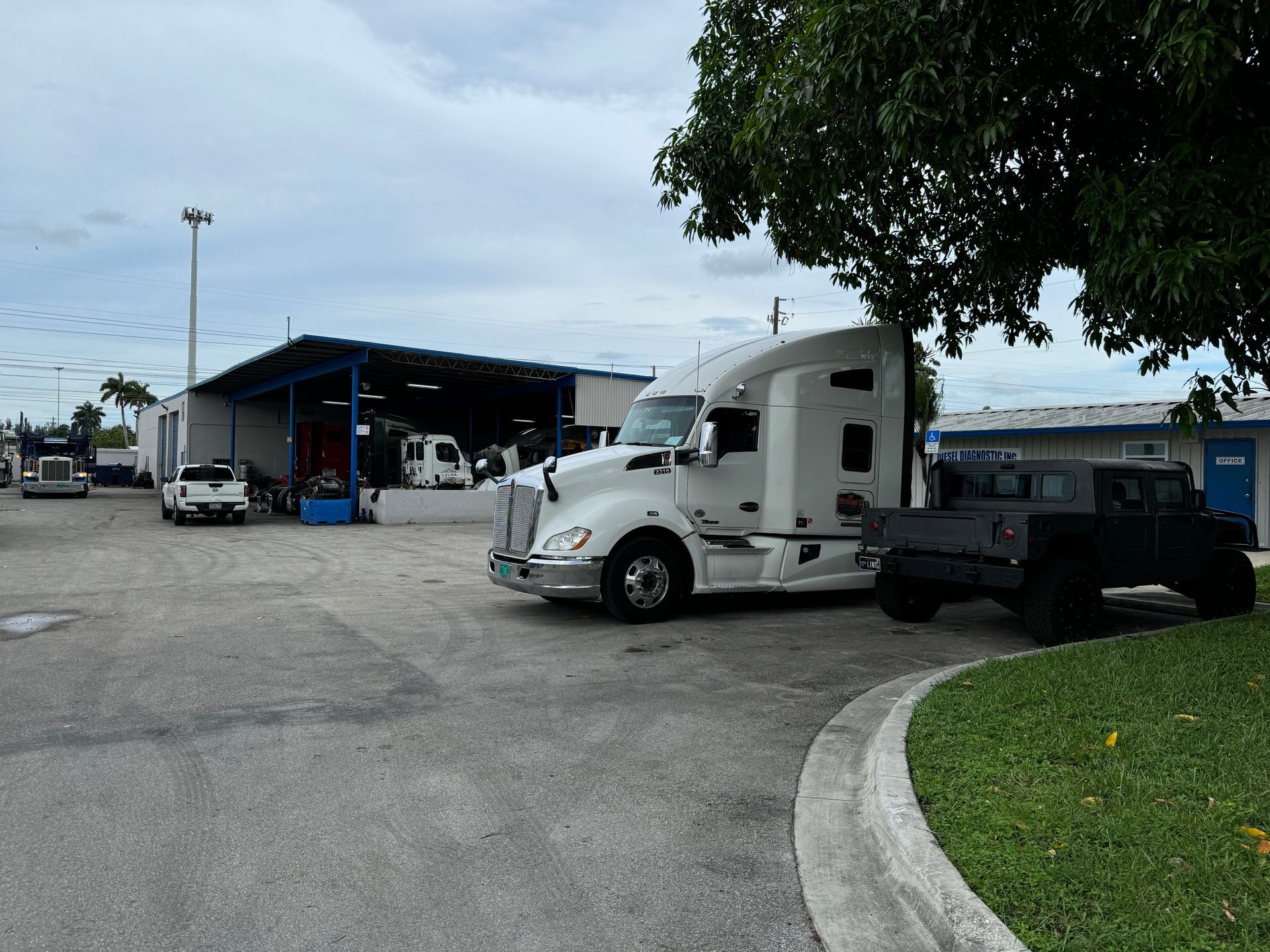 A white semi-truck parked in a gravel lot next to a black Humvee, with a maintenance garage and trees in the background.