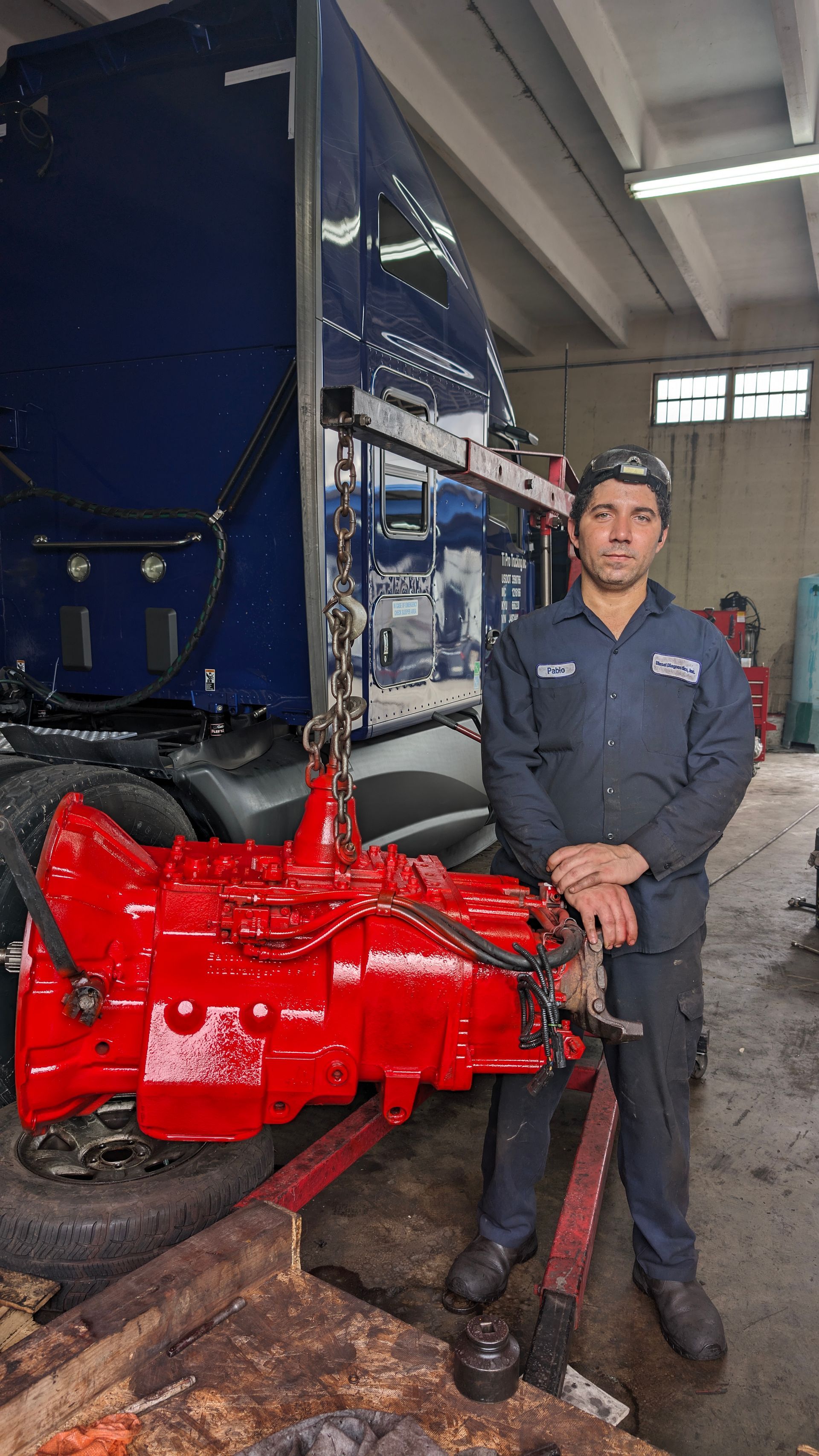 A mechanic stands in a shop next to a large, newly painted bright red vehicle transmission hanging from a hoist.