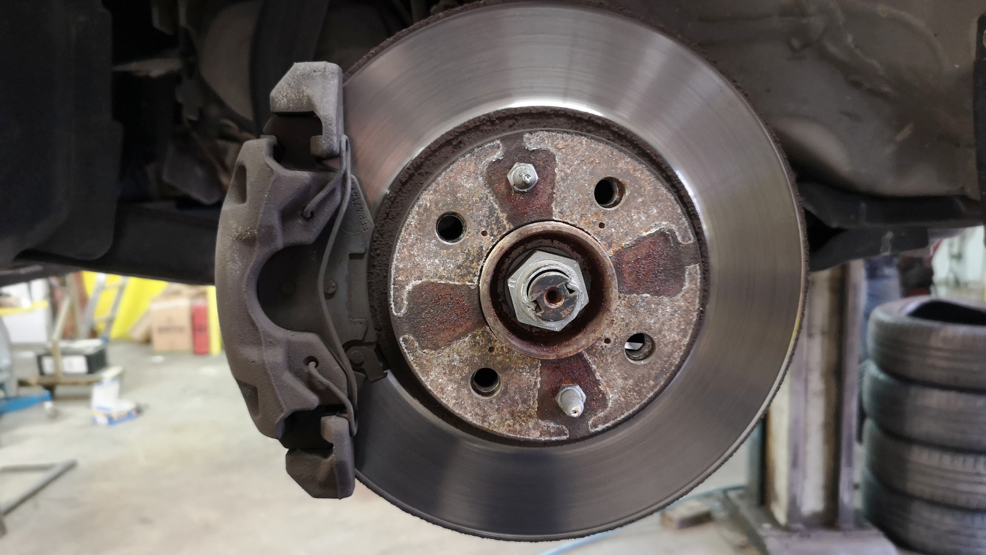 A close-up of a car's disc brake rotor and brake caliper assembly mounted on a wheel hub inside an auto repair shop.