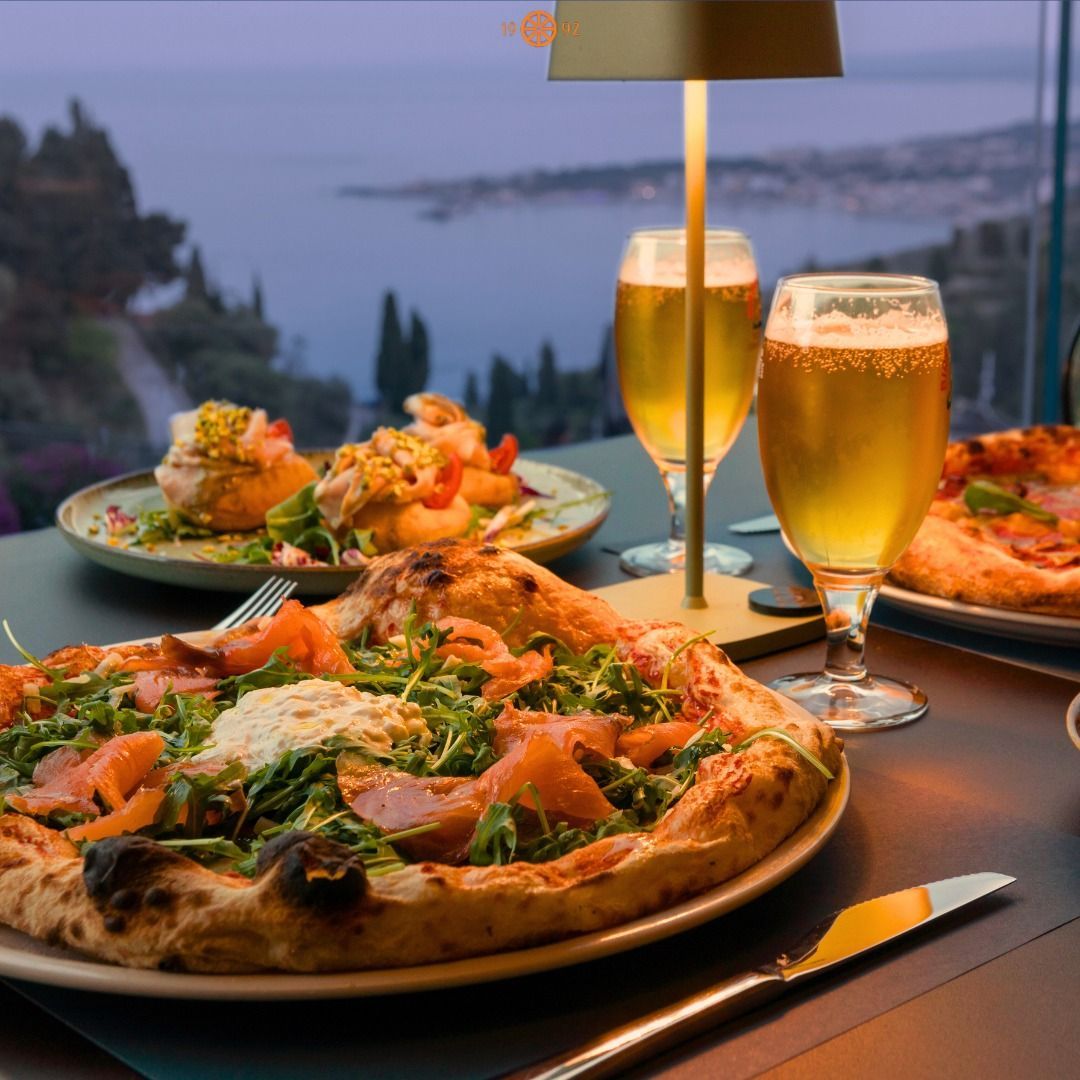 Una pizza, degli antipasti e birre su un tavolo con vista sul paesaggio costiero al tramonto.