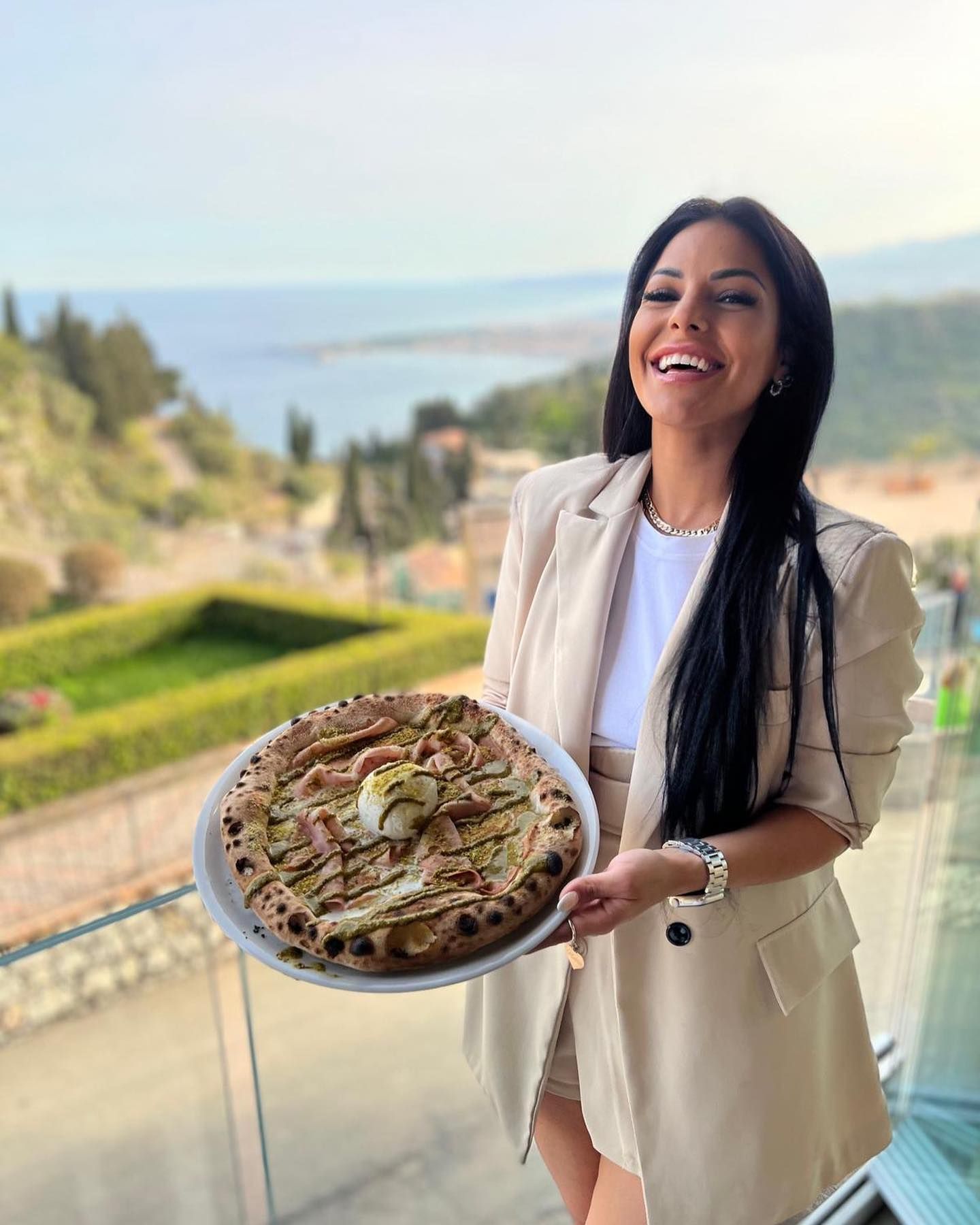 Donna sorridente con in mano una pizza sul balcone con vista sul mare.