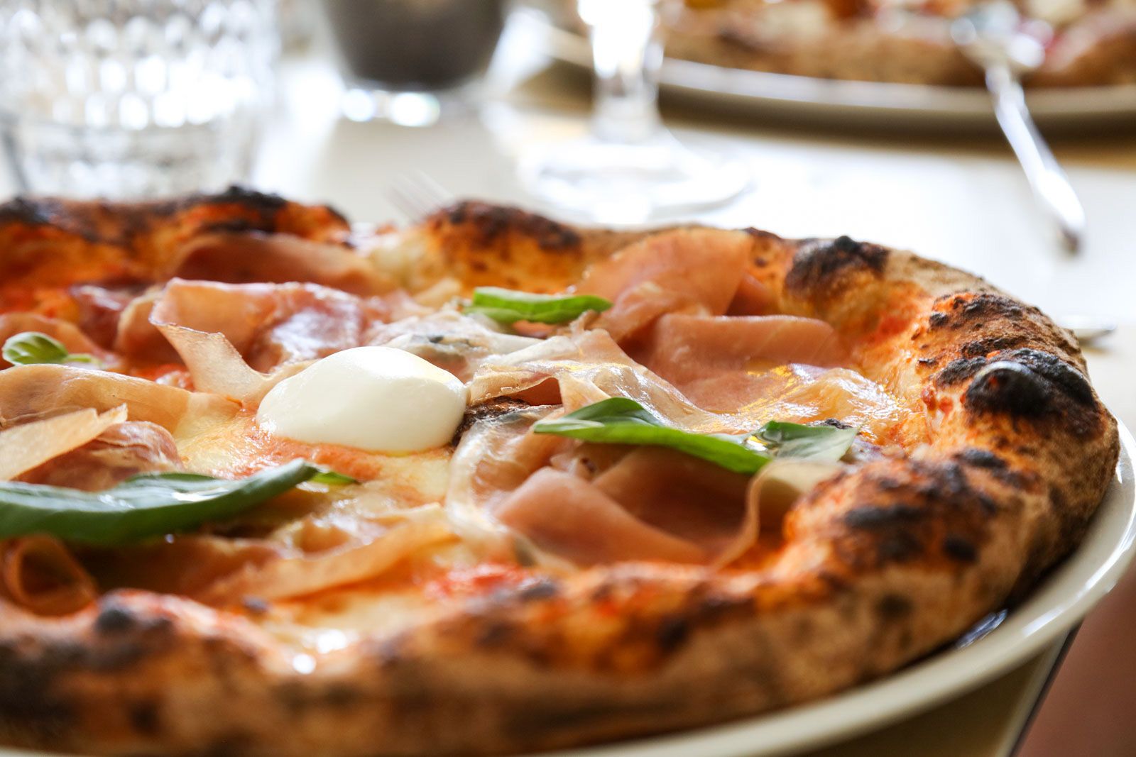 Primo piano di una pizza con condimenti tra cui prosciutto, formaggio e basilico su un piatto bianco.