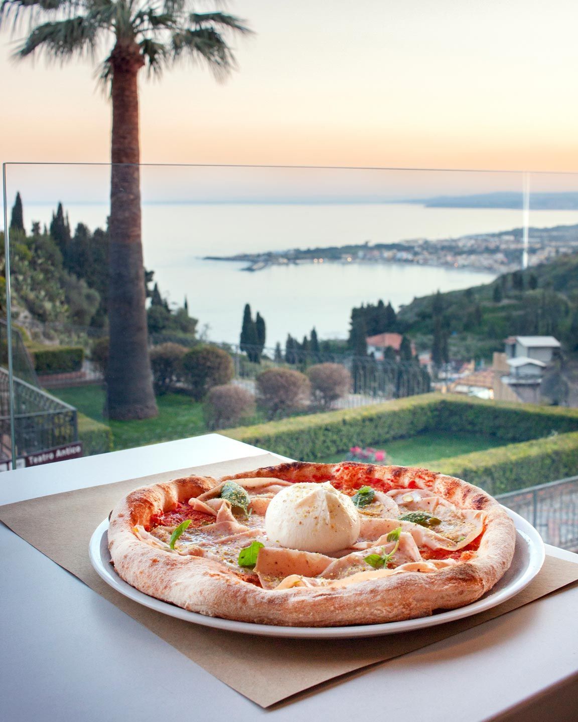 Pizza condita con burrata su un piatto, con vista panoramica sull'oceano sullo sfondo.